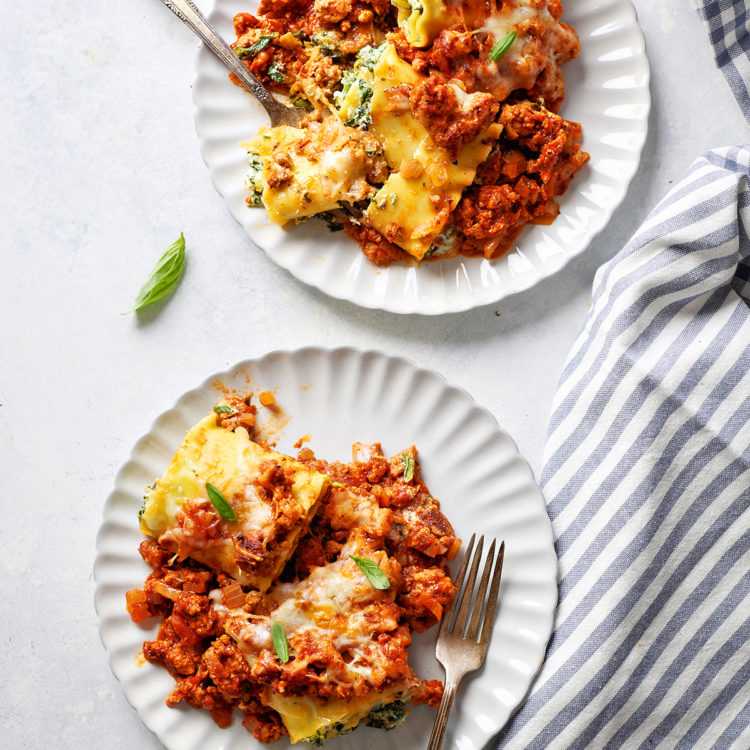 Two plates of sausage lasagna rolls served on white plates with forks, garnished with fresh basil. A bite has been taken from one piece, revealing the cheesy spinach filling.