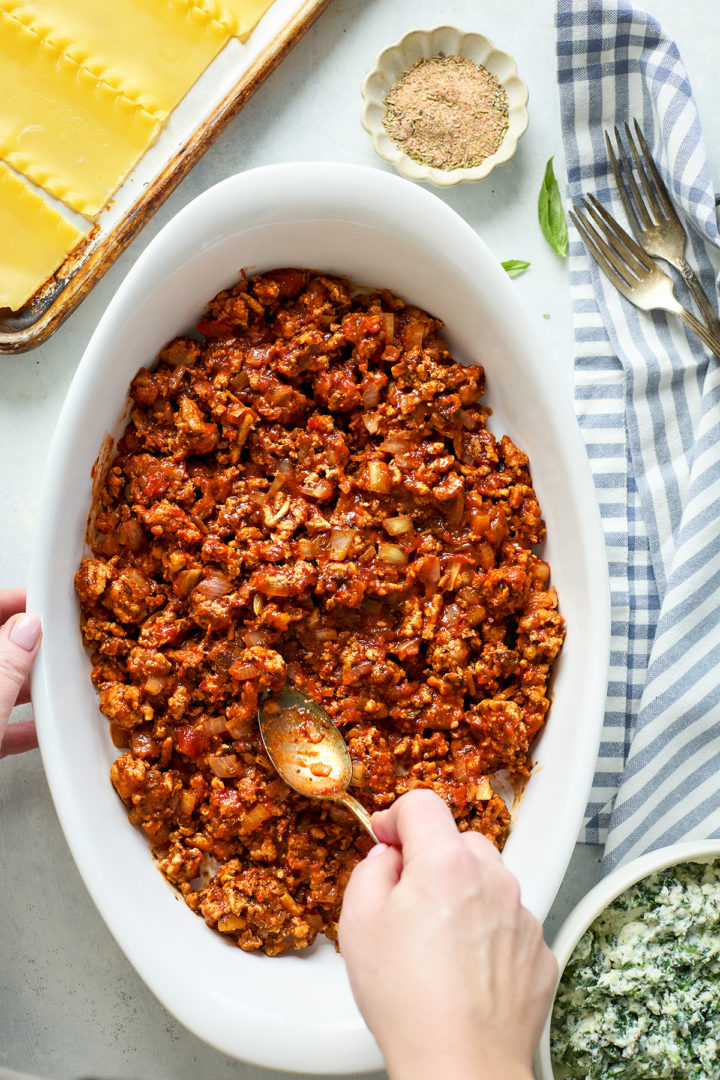 Overhead shot of a woman adding homemade meat sauce to a white baking dish, with bowls of meat sauce and spinach ricotta filling, a blue plaid kitchen towel, forks, fresh basil leaves, and a small bowl of seasoning in the background