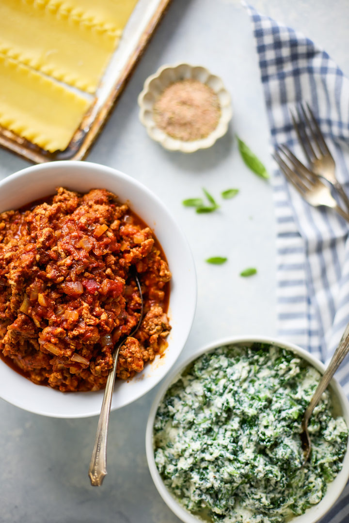 Overhead shot of bowls filled with meat sauce and spinach ricotta filling, surrounded by a blue plaid kitchen towel, forks, fresh basil leaves, a small bowl of seasoning, and a baking pan with cooked lasagna noodles