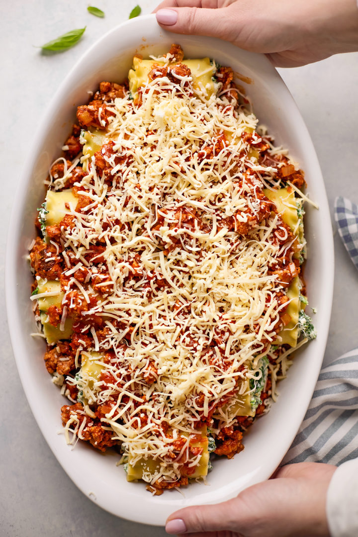 Woman holding a white baking dish filled with sausage lasagna rolls, topped with a rich, meaty marinara sauce and shredded mozzarella and parmesan cheese, ready to bake.