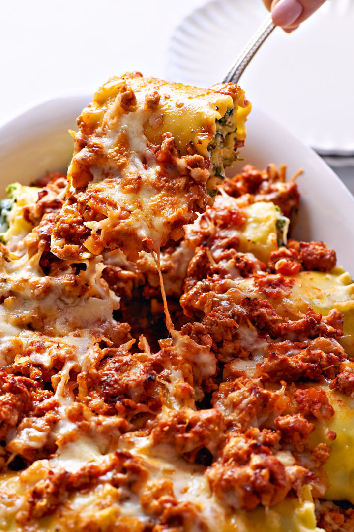 A woman serving cheesy sausage lasagna rolls from a white baking dish using a metal serving utensil. The dish is topped with melted cheese and a rich, meaty sausage sauce