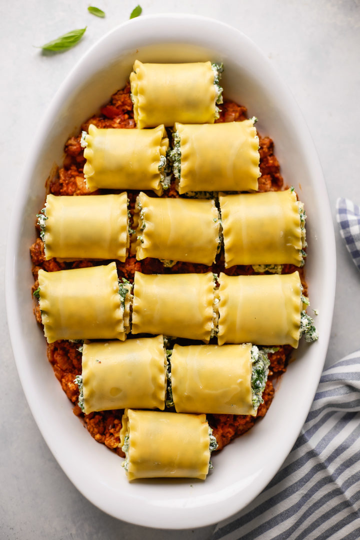 Lasagna rolled up and placed on top of a layer of savory sausage meat sauce in a white baking dish, ready for baking.