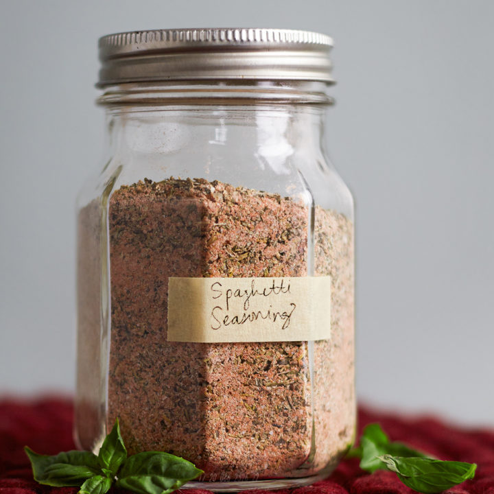 A glass jar filled with homemade spaghetti seasoning sits on a red felt trivet atop a rustic wooden table surrounded by fresh basil leaves