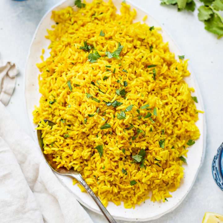 Turmeric yellow rice on an oval white platter, with fresh cilantro and a kitchen towel nearby. S spoon rests on the plate, ready to serve.