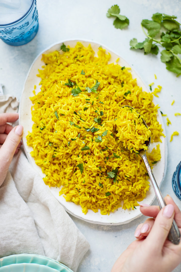 A woman serving turmeric rice recipe from a white platter, surrounded by fresh herbs, serving plates, and a kitchen towel for a cozy presentation.